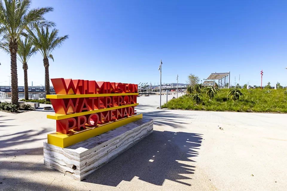 Person-height red 'Wilmington Waterfront Promenade' sign on concrete walkway with palm trees in background on waterfront walkway at the Port of Los Angeles, California.